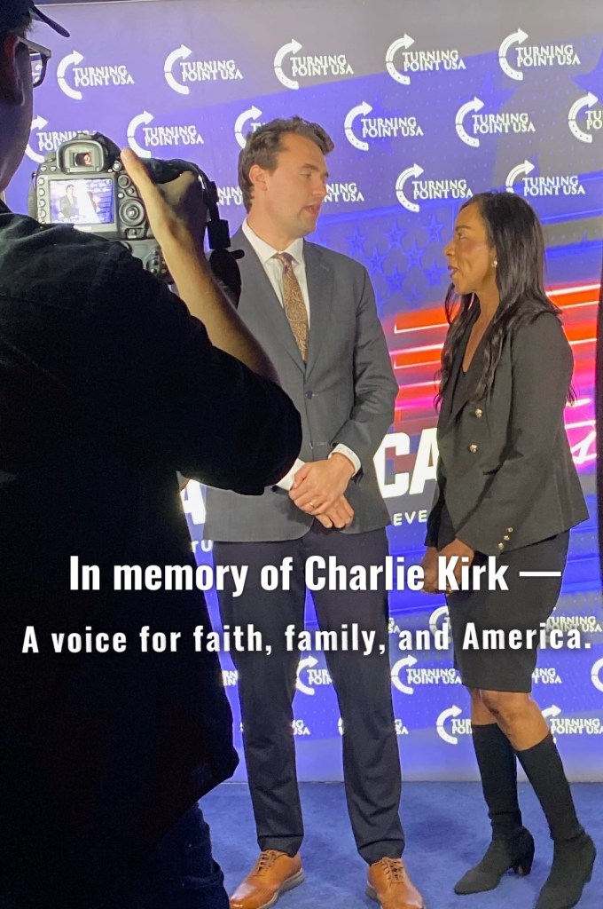Two individuals engaged in a conversation at an event, with a backdrop featuring the Turning Point USA logo and a tribute caption to Charlie Kirk.